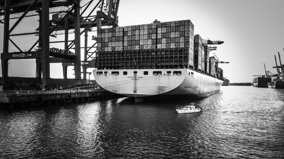 Grayscale image of a cargo ship at port, illustrating the scale of global maritime shipping vulnerable to disruption