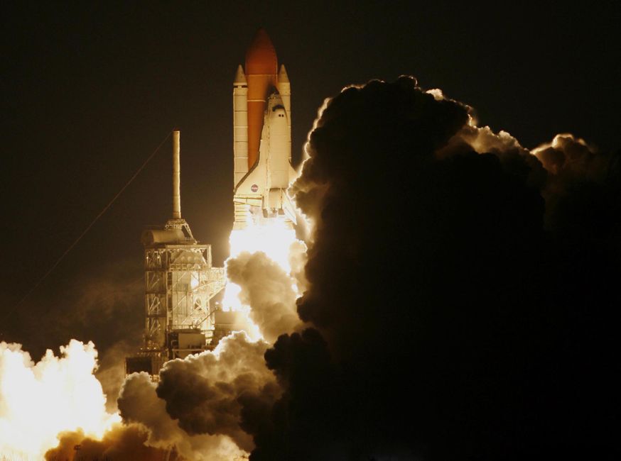 A rocket launches into the night sky, shrouded in smoke and illuminated by its exhaust flame.