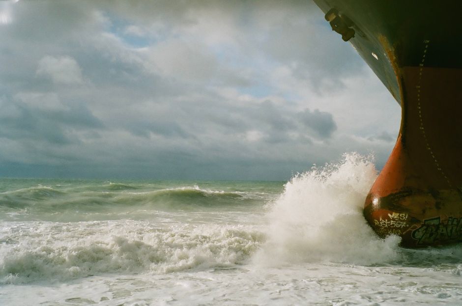 A ship's bow cutting through ocean waves under a cloudy sky, representing maritime commerce through contested waters.