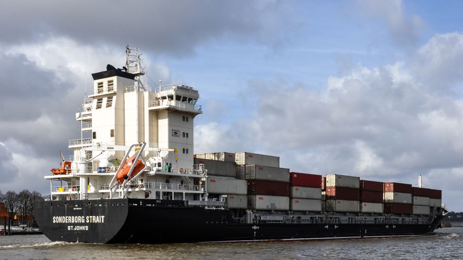 Container ship navigating harbor with stacked cargo under a cloudy sky