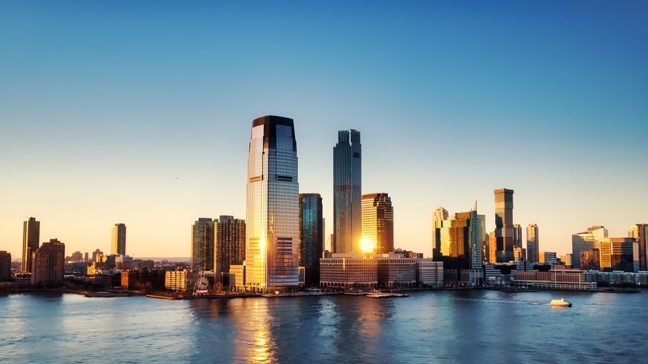 Sunrise reflecting off glass skyscrapers in a major city skyline, viewed from across the water.