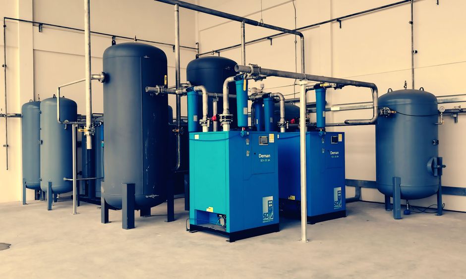 Industrial air compressor system with blue tanks and pipes inside a manufacturing plant, representing semiconductor fabrication equipment.