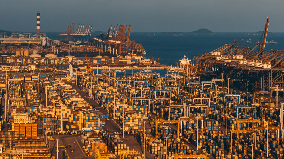 Aerial view of a bustling commercial port with stacked shipping containers and cranes along a waterway.