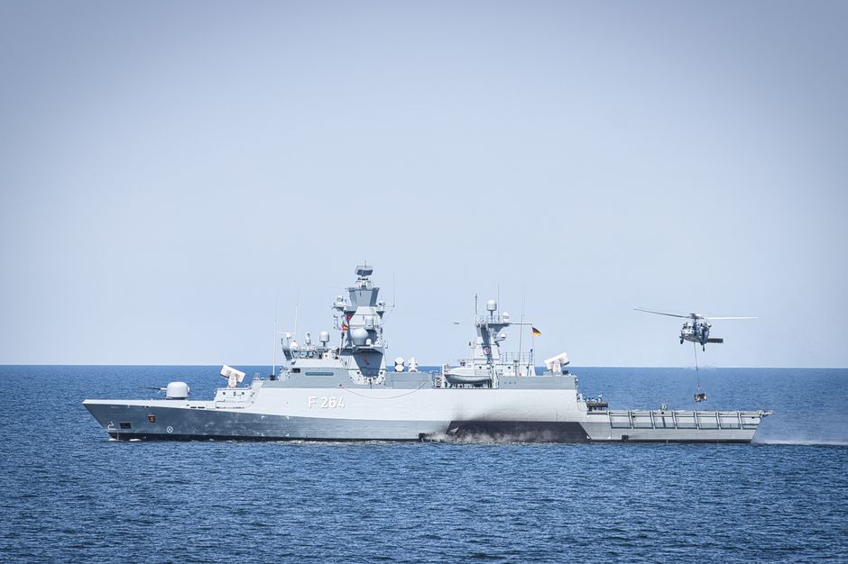A naval frigate navigates open waters with a helicopter hovering above, illustrating maritime security operations in a contested shipping lane.