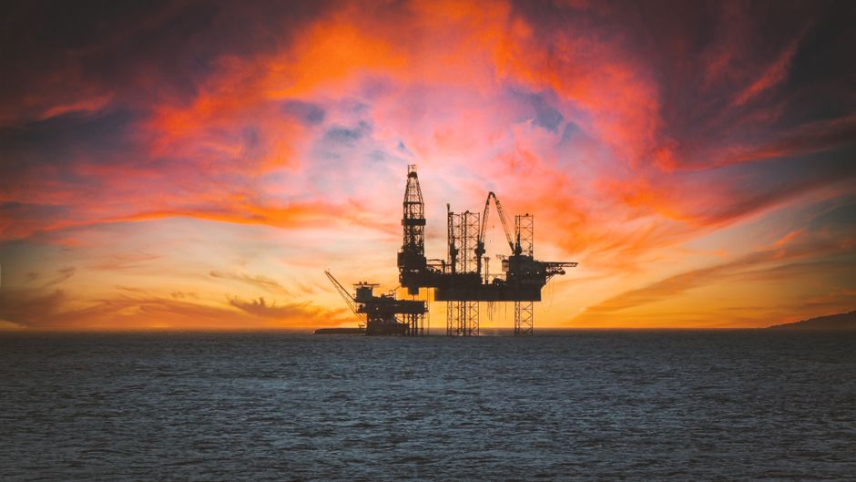 Offshore oil platform silhouetted against a dramatic sunset sky over the sea.