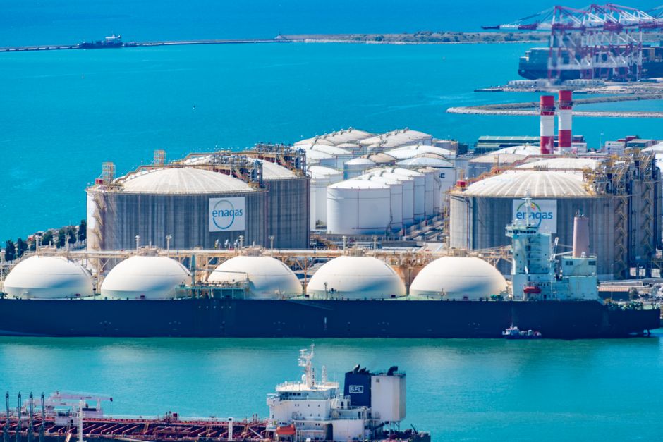 Aerial view of an LNG gas terminal with storage tanks and tanker ships in turquoise waters.