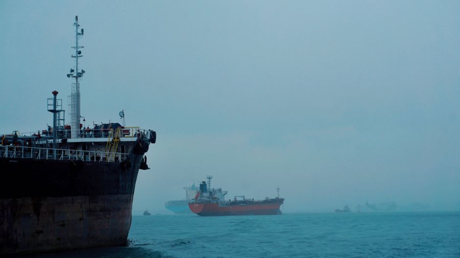 Cargo ships and an oil tanker move through a hazy sea as geopolitical disruption raises shipping and energy risks.