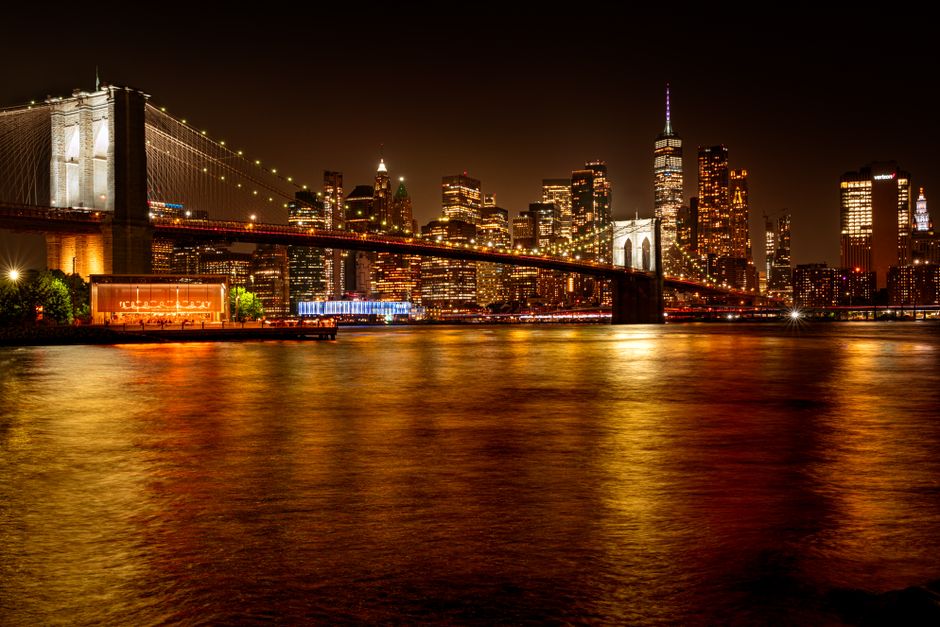 Nighttime view of the Brooklyn Bridge and Manhattan skyline with city lights reflecting on the East River.