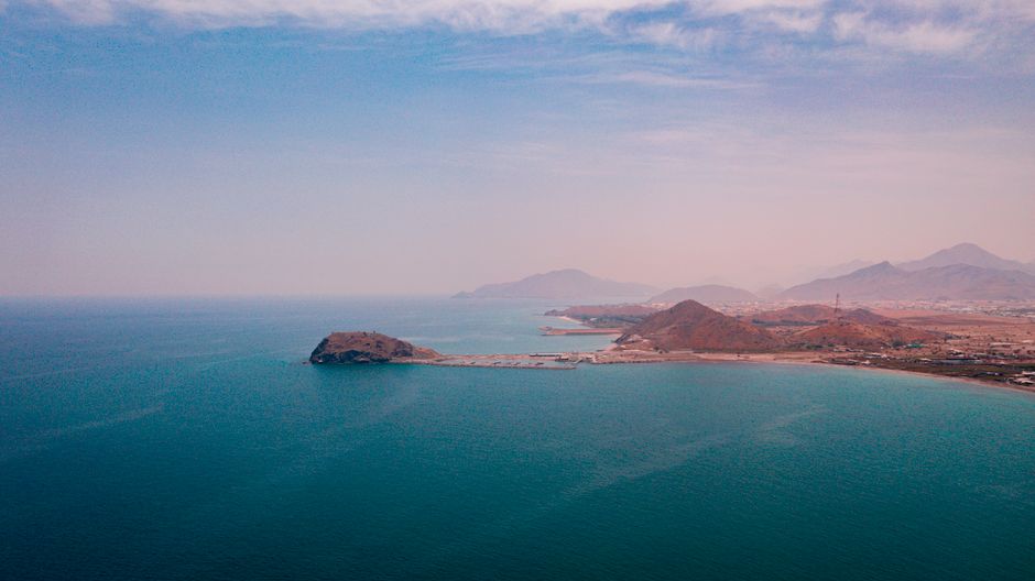 Aerial view of a coastal settlement beside a mountain range and open ocean, evoking strategic maritime geography.