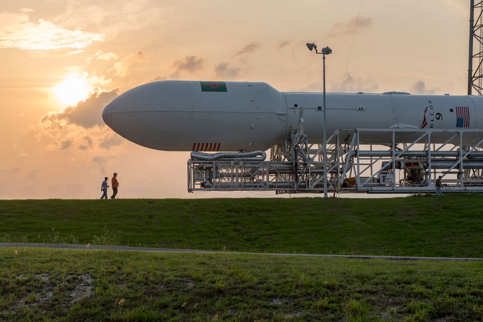 A rocket on a launchpad at sunset with people standing nearby, silhouetted against the sky.