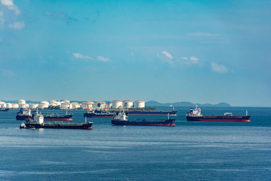 A fleet of cargo ships docked near oil storage tanks along a serene coastline with a clear blue sky, illustrating the shipping chokepoint crisis in the Red Sea and Strait of Hormuz.