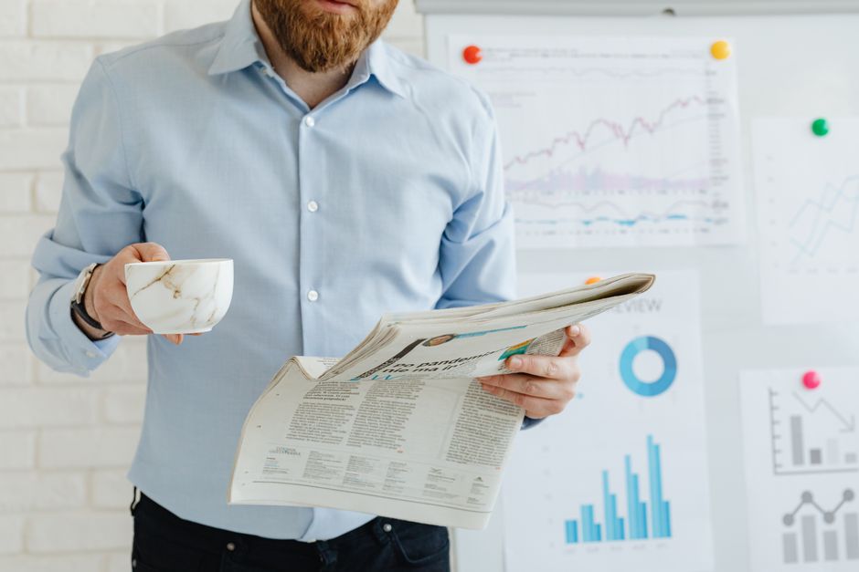 Business person holding a newspaper and coffee in an office with financial charts