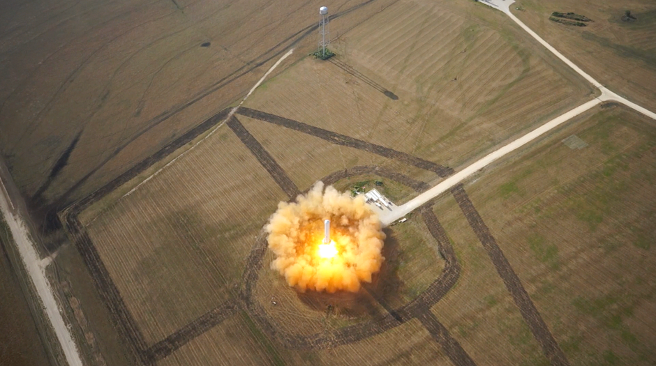 Aerial view of a rocket lifting off from a launchpad in open terrain, trailing exhaust plume.