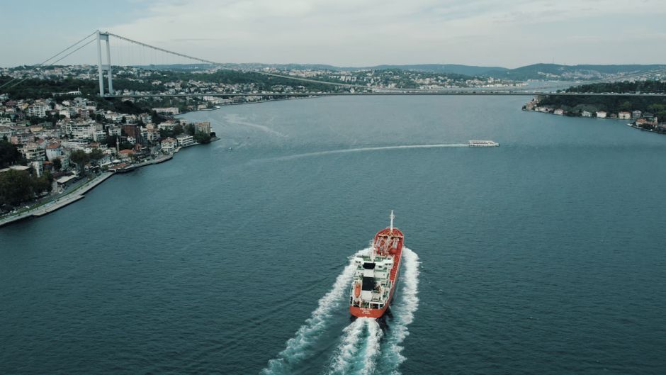 Aerial view of a large cargo ship navigating a narrow strait beneath a bridge, illustrating the critical maritime chokepoint at the Strait of Hormuz where one-fifth of global oil supplies once flowed.