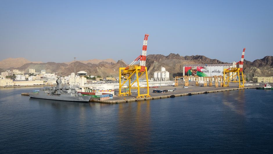 Aerial view of Muscat port with a naval ship, cranes, and mountains in Oman.