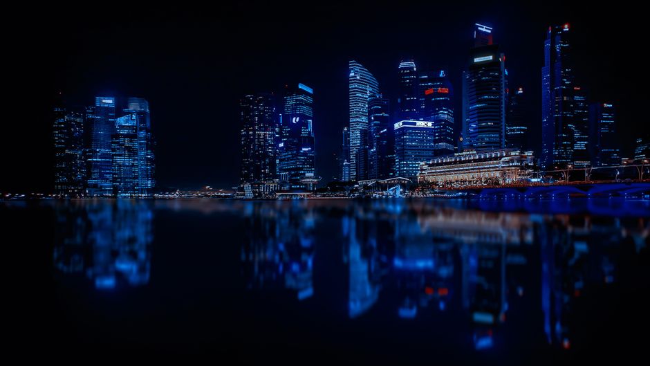 Nighttime aerial view of a city skyline with illuminated skyscrapers reflecting on calm water.