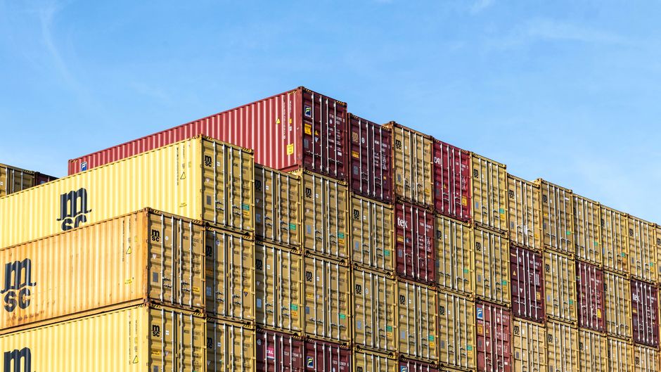 Stacked cargo containers at a port terminal under a clear sky, illustrating the global shipping and trade infrastructure now disrupted by the Hormuz crisis.