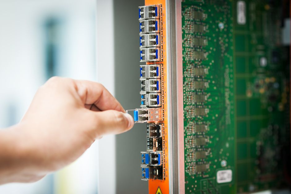 An engineer connecting a cable into a network server port in a data center room, with rows of server racks in the background.