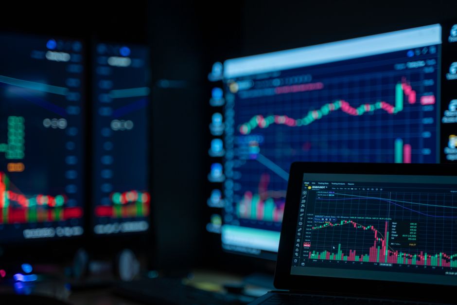 Multiple monitors displaying stock market charts and financial data in a dimly lit trading room.