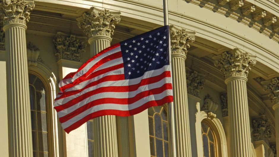 US flag waving on a pole outside the US Capitol Building in Washington, DC, with the dome visible in the background.