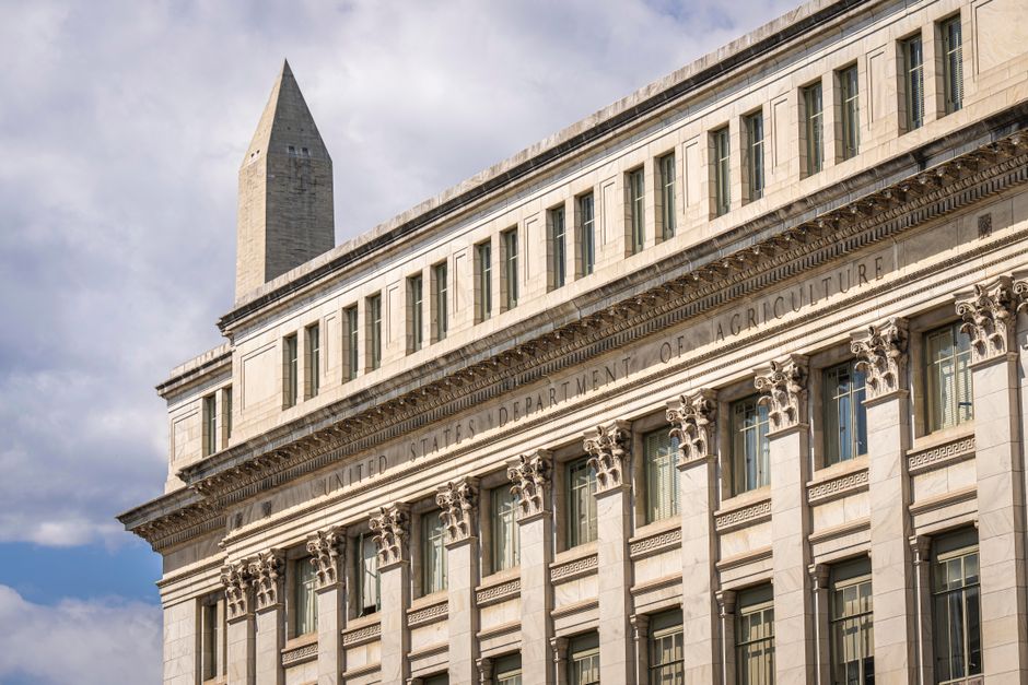 US government building with the Washington Monument visible behind it under a cloudy sky