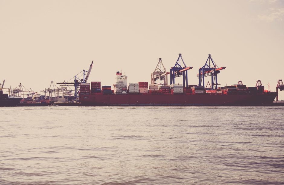 A cargo ship loaded with containers docked at a seaport with cranes, illustrating global maritime shipping and energy trade infrastructure.