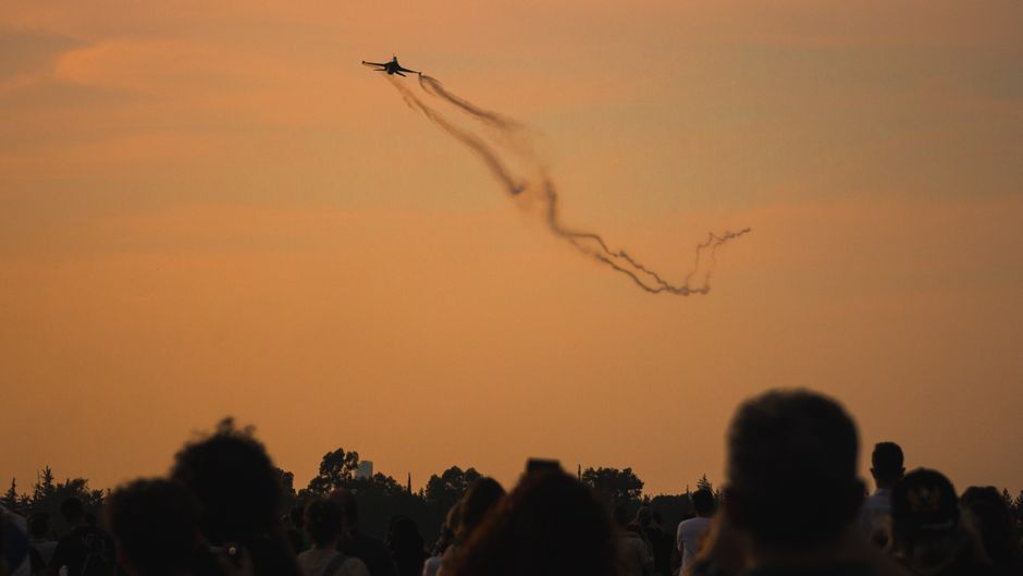 A military jet flies over a crowd at an airshow, leaving trails against a warm sunset sky.