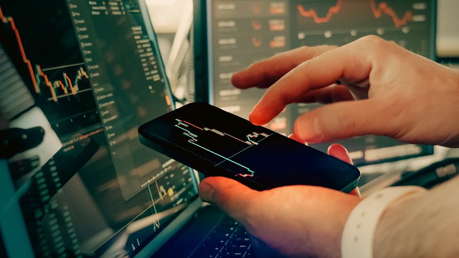 Hands interact with a smartphone displaying financial trading data on digital screens.