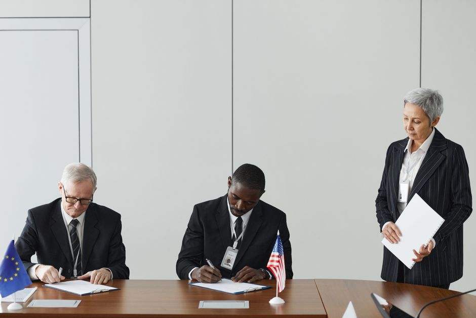 Executives signing an international agreement with EU and US flags displayed on a wooden table.