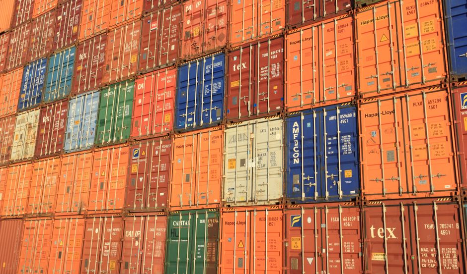 Stacked shipping containers represent the freight and tariff channel into home-goods costs