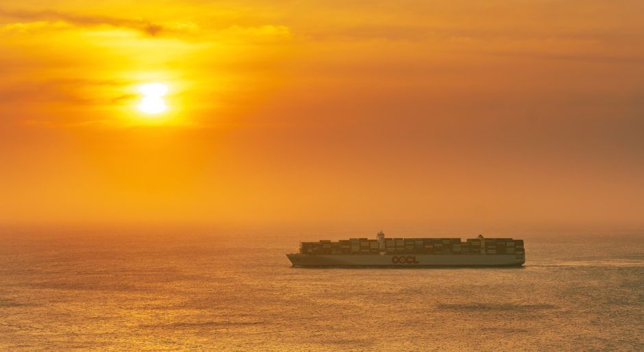 A large cargo container ship sailing across open ocean under a vibrant orange sunset, representing global maritime supply chains.