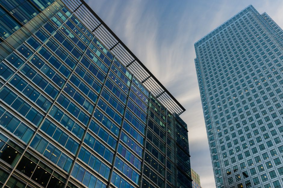 Upward perspective of modern glass skyscrapers against a blue sky, representing financial district corporate headquarters and capital markets.