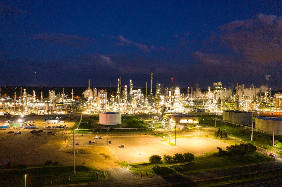 Aerial view of an illuminated oil refinery at night, with industrial structures glowing against the dark sky.
