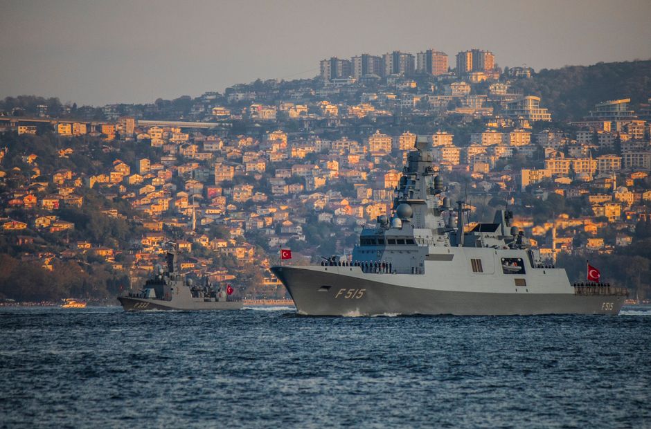 Naval warships sailing through a narrow strait with hillside buildings visible along the shore.