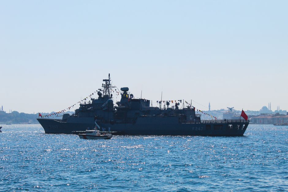 Naval vessel transiting a strategic waterway near a coastal city.