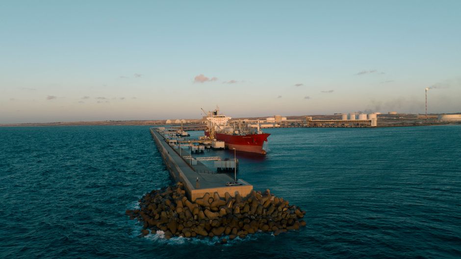 A cargo ship sits at an industrial port, representing the strategic shipping risk around the Strait of Hormuz.