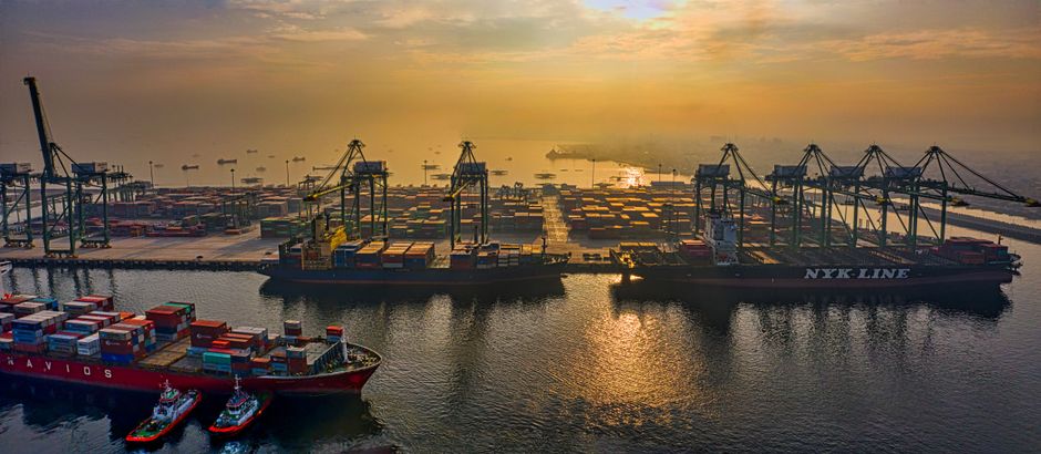 Aerial view of a bustling container port at sunset, illustrating the vulnerability of global shipping infrastructure to geopolitical disruption.