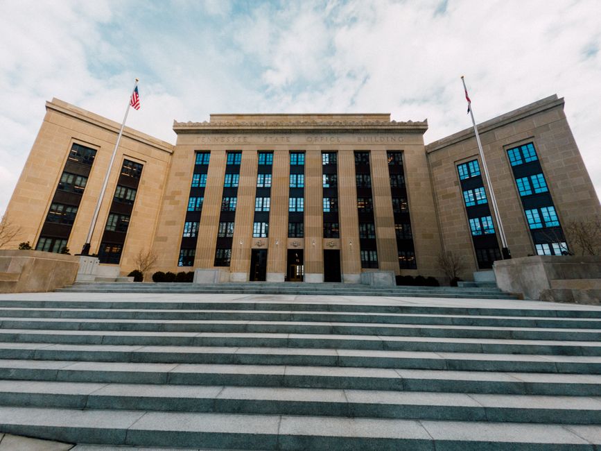 From below, a federal building exterior with American flags and a wide staircase under a cloudy sky.