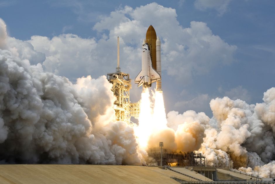 A rocket lifts off in a column of smoke against a bright sky