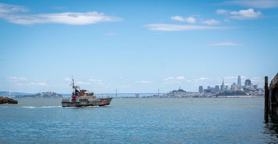 A naval warship patrolling coastal waters with bridges and skyline in the background, illustrating the military blockade of the Strait of Hormuz and Bab al-Mandeb chokepoints