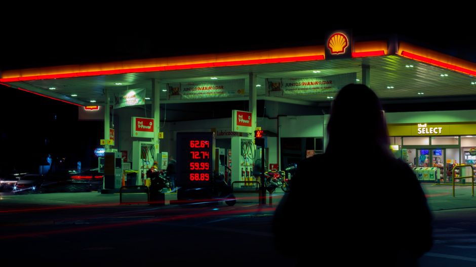 A gas station illuminated at night with fuel pumps visible under overhead lighting, a person's silhouette in the foreground.