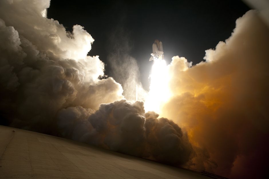 A space shuttle launching into the night sky, surrounded by billowing smoke and intense light.
