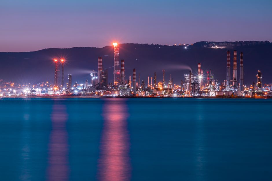 Aerial view of an oil refinery industrial complex at twilight with illuminated facilities reflecting on water.
