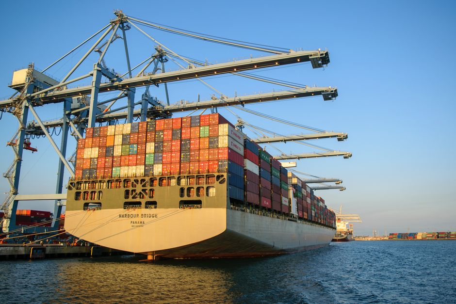 Large cargo container ship docked at an industrial port with loading cranes under a clear blue sky.
